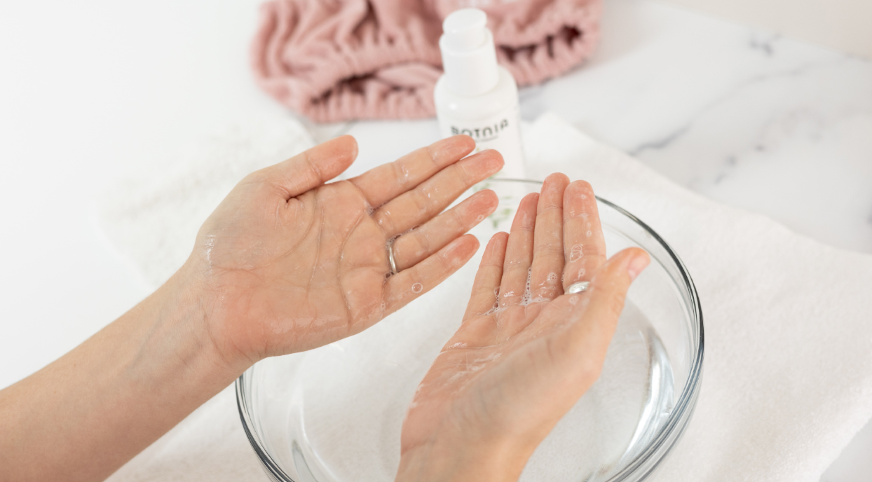 A pair of hands above a bowl of water, a pink Tassi hairband, and a bottle of Botnia Cleanser.